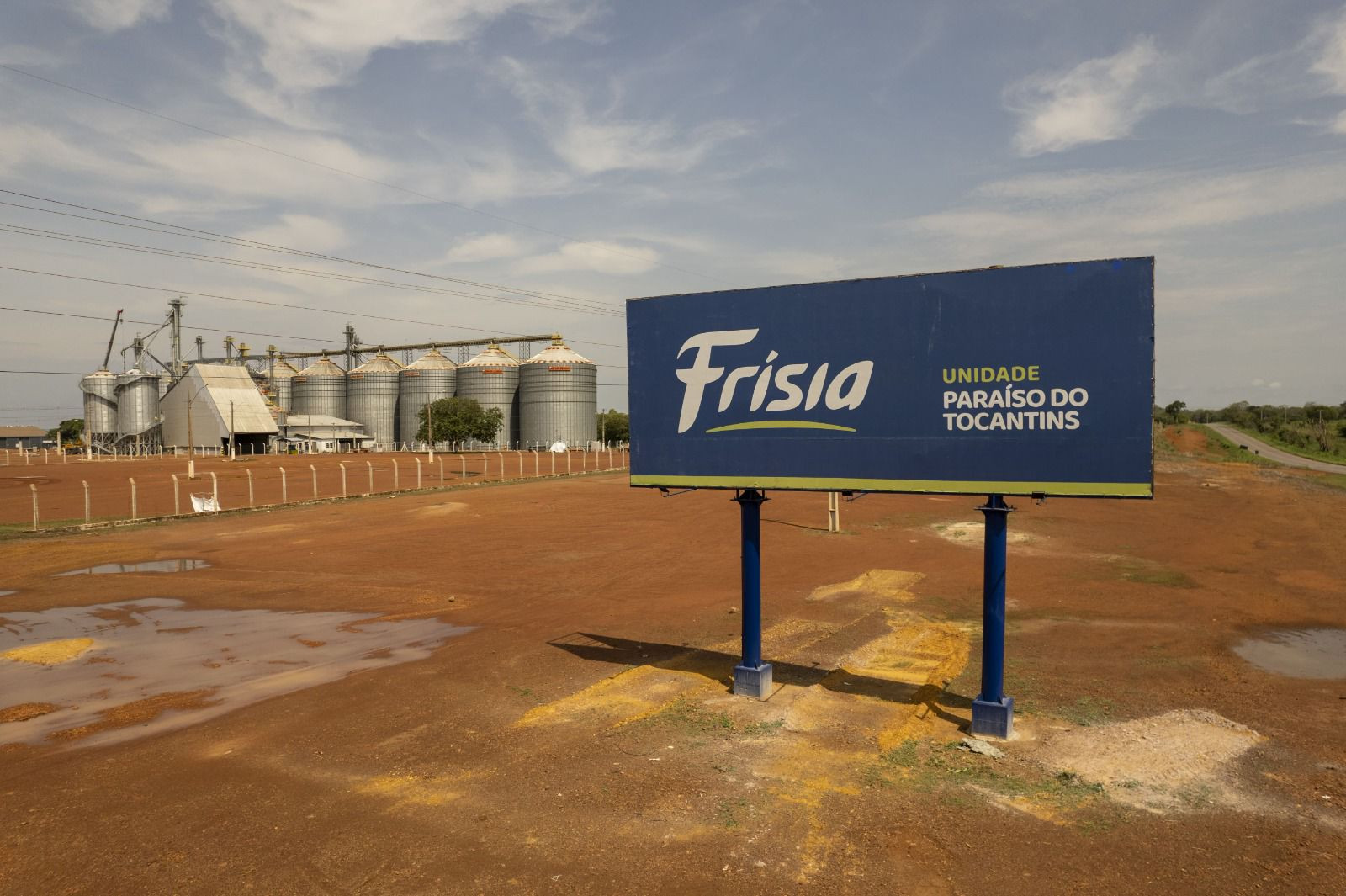 A imagem mostra a unidade da Frísia em Paraíso do Tocantins, com uma placa azul em primeiro plano e, ao fundo, silos metálicos de armazenamento de grãos em uma área rural de solo avermelhado.
