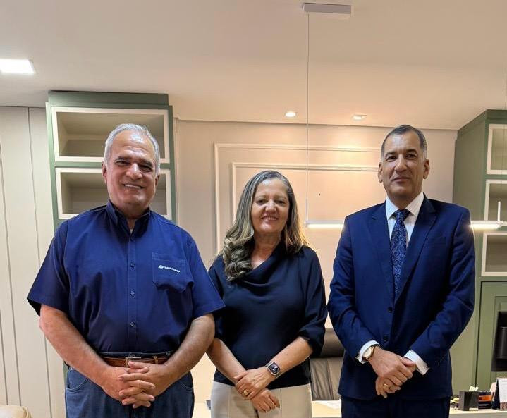 A imagem mostra três pessoas em um ambiente de escritório. Duas delas são homens e uma é mulher. O homem à esquerda está vestido com uma camisa azul escura e calça jeans, sorrindo. A mulher no meio usa uma blusa azul escura e calça clara, também sorrindo. O homem à direita veste terno azul escuro com gravata e tem expressão séria. Todos estão em pé, lado a lado, olhando para a câmera. O ambiente ao redor é claro e organizado, com móveis de escritório ao fundo.